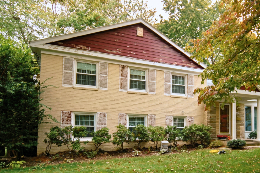 A two-story tan brick house with dark red gables showing significant fading paint and wear, surrounded by green trees and a leafy lawn.